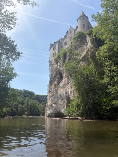 The image presents a view of Walzin Castle, situated in Dinant, Belgium. The castle perches atop a rocky cliff face. The River Lesse flows below the castle and cliff, its surface reflecting the environment. The cliff face is partly covered with vegetation. Trees line the riverbank on the opposite side of the cliff. The sky is blue with contrails visible. Kayaks and people are visible on the River Lesse.