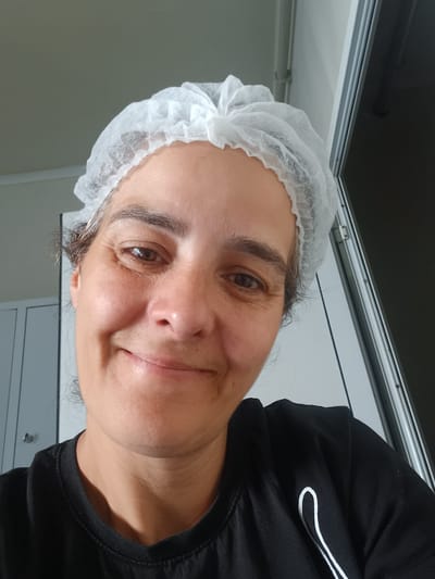 A person is shown in a close-up, sideways selfie, wearing a black t-shirt and a white disposable hair net. The person is smiling slightly at the camera, with their head tilted. Their eyes are brown and they have dark eyebrows. The background is blurred, but appears to be an indoor setting with neutral-colored walls and cabinets. The lighting suggests it is daytime. There is no visible text in the image. The context provided indicates the location is Rakevo, Bulgaria.