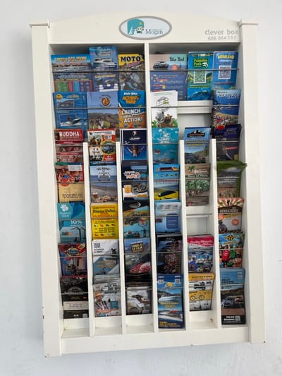 A white, vertical, wooden rack holds rows of brochures. The brochures advertise various tourism-related activities and services. The rack is located in a building in Mogan, a town on the southwestern coast of Gran Canaria, Spain. The brochures are arranged in rows of eight columns. The rack has a sign at the top that reads "clever box 606 064 257." The brochures depict activities such as water sports, boat trips, sightseeing, and jeep safaris. Some of the brochures are in Spanish and English. A few of the brochures have QR codes on them. The rack is attached to a white wall.