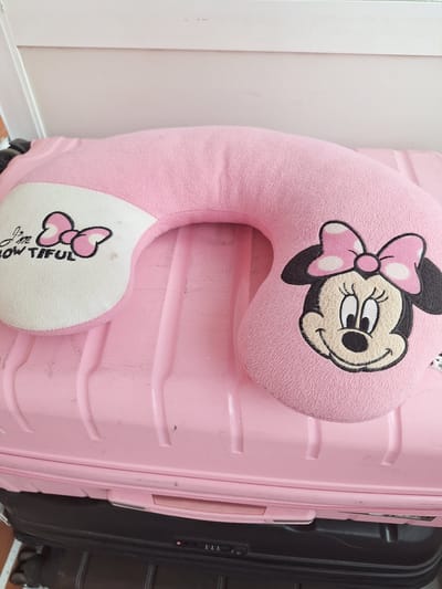 The image shows a pink neck pillow with a Minnie Mouse design, placed on top of a pink suitcase. The suitcase has a textured surface with horizontal grooves. To the right of the pink suitcase, a portion of a dark-colored suitcase is visible. The background consists of a plain, light-colored wall and a white baseboard. The pillow features a close-up of Minnie Mouse's face with her signature bow, and a smaller bow design with the text "I'm bow tiful" on the bottom curve of the pillow. The overall scene appears to be indoors, possibly in a room or hallway. There is no indication of the time of day, weather, or specific emotions. The location context provided is Vologda, Russia.