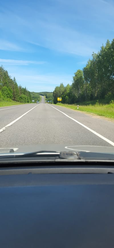 The image shows a view from inside a car driving on a two-lane asphalt road in the countryside. The road is lined with trees on both sides. The sky is blue with some scattered clouds. There are a few cars in the distance on the road. On the right side of the road, there is a yellow sign with some text and numbers, partially obscured by trees. The foreground shows the dashboard and windshield of the car. The location is identified as Крэўскі сельскі Савет, Belarus.