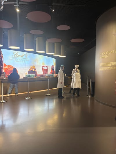 The image shows the interior of the Lindt Home of Chocolate museum in Kilchberg, Zurich, Switzerland. Two staff members wearing white coats and hats stand near a display screen featuring the Lindt logo and images of chocolate. A barrier is set up to guide visitors near the screen. The wall behind the display features a dark backdrop with cylindrical lights and round pink decorations. Above the staff is text that reads "CHOCOLATE HEAVEN". The area appears to be a visitor attraction within the museum.