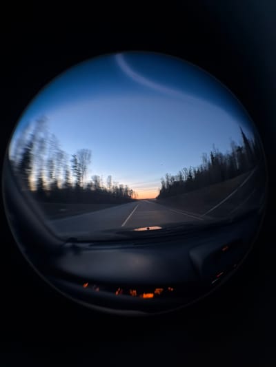 The image appears to be a reflection in a convex mirror or lens, presenting a distorted view of a highway at twilight. The highway has a central yellow line. Trees line the side of the road. There are reflections of an interior car dashboard. The environment is generally dark, suggesting dim lighting or night.