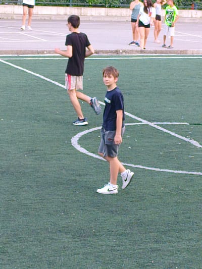 The media file shows a group of people on a grassy field with white lines, likely a sports field. In the foreground, a young boy with brown hair wears a dark blue t-shirt and gray shorts, with white athletic shoes. He is walking and looking to his left, with a neutral expression. Behind him, another boy in a black t-shirt and light-colored shorts with red stripes is also walking, his gaze directed forward. He is wearing dark athletic shoes. In the background, several other individuals are visible, some standing and some walking. One person in a bright green t-shirt and white shorts stands out. The lighting suggests it is daytime, possibly late afternoon or early evening, and the weather appears to be fair. The setting is an outdoor sports facility, with a track or paved area visible behind the field. The overall scene appears to be casual and relates to a sporting or recreational activity. No visible text is present in the image.