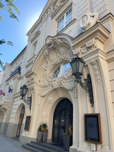 The image shows the facade of a building in Bratislava, Slovakia, likely a historical building due to its ornate architectural style. The building is light-colored, possibly sandstone or stucco, and features intricate decorative elements such as carvings, garlands, and urns. The main entrance is a large, arched doorway with decorative black metalwork. Flanking the entrance are large, wrought-iron lamps. Two flags are visible on the left side of the building, one of which appears to be the Slovak flag. A sign is mounted on the wall to the right of the entrance, displaying text and graphics, possibly indicating an exhibition or event. There are planters with greenery near the entrance. The sky is bright blue, suggesting a clear day, with some branches of a tree visible in the upper left corner. The perspective is from a low angle, looking up at the building.