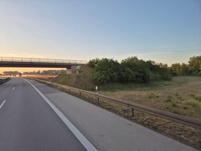 The image shows a highway overpass with a pedestrian walkway crossing over the road. The highway is curving to the left and appears to be in a rural or semi-rural setting. The sky is clear and blue with a faint glow of sunrise on the horizon. Trees and grassy areas are visible on either side of the highway. A metal guardrail runs along the side of the highway. A black and white pole with a reflective marker is visible on the side of the road, likely a road marker. The scene is set during early morning, as suggested by the soft light and the mist in the distance. The location is identified as Altötting, Germany.