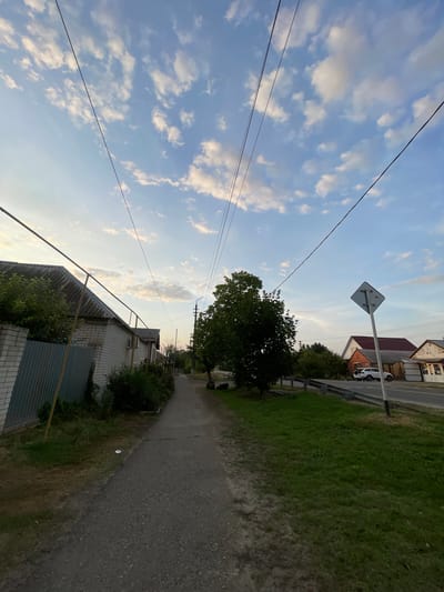 The image depicts a paved sidewalk running alongside a street in the city of Donskoye, Russia. On the left, a white brick house with a gray corrugated metal fence is visible. On the right, a grassy area borders the street, which is lined with more houses and trees. Power lines stretch across the sky, and a diamond-shaped road sign stands near the right edge of the frame. The sky is blue with scattered white clouds, suggesting it is daytime. The scene appears to be a quiet residential area.