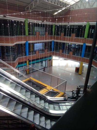Captured from an elevated perspective, the image depicts the interior of a multi-level structure, likely a shopping mall or commercial building, in Parroquia Guatire, Venezuela. The building features brick walls, with windowed storefronts on multiple levels. Festive white string lights are strung across the open atrium, vertically descending between floors. Vertical supports are alternately painted blue and green.
An escalator dominates the foreground. Below, a rectangular trampoline with yellow padding is visible on the main floor.
A lone individual is walking across the main floor.
The environment appears relatively empty and brightly lit, possibly by natural light from the roof. The overall scene suggests a commercial setting that is somewhat underutilized at the time of the photograph.