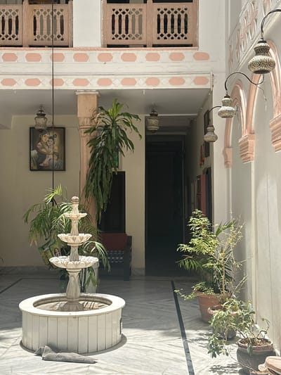 This is an outdoor courtyard in Pushkar, India, featuring a traditional Indian architectural style. In the foreground, a multi-tiered white marble fountain is centrally positioned on a marble-tiled floor. To the left of the fountain, a painting depicting a mother and child, possibly a religious figure, hangs on a light-colored wall. Several potted plants, including palm trees and a spindly plant with small leaves, are arranged throughout the courtyard. Decorative mosaic lanterns hang from the ceiling on curved metal fixtures. The courtyard is enclosed by walls with arched doorways and decorative railings on the upper levels. Sunlight casts shadows across the floor, indicating a daytime setting. A folded gray towel lies on the floor in front of the fountain.