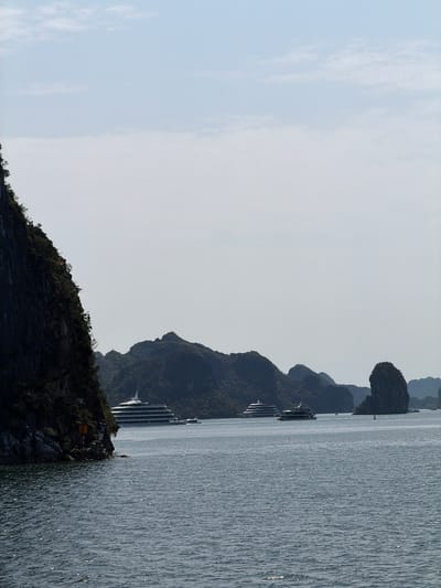 The image depicts a panoramic view of a scenic bay, characteristic of Ha Long Bay, Vietnam, featuring numerous limestone karst islands and mountains rising from the water. The sky, occupying the upper portion of the frame, is bright and hazy with some faint, wispy clouds.
Across the mid-ground, a chain of dark, vegetated islands and mountains stretches horizontally, appearing as silhouettes against the lighter sky. Several large, multi-deck cruise ships or tour boats are visible on the calm water, interspersed among these islands. These vessels are predominantly white with dark window detailing, indicating their use for tourism.
The lower part of the image shows the surface of the bay, which is covered in small ripples and glitters with countless reflections of sunlight, creating a shimmering effect. In the lower-left foreground, a dark, rocky island with sparse vegetation is partially visible. On this island, there are two small signs: a white one featuring a blue symbol or logo (possibly a flag or local marker) and a yellow sign below it, the text of which is unreadable.
The scene takes place during bright daylight, likely late morning or early afternoon, as suggested by the strong sun reflections on the water and the clear conditions. The weather appears pleasant and clear. No individuals are distinctly visible, but the presence of multiple large boats implies active tourism or leisure cruising in the bay.