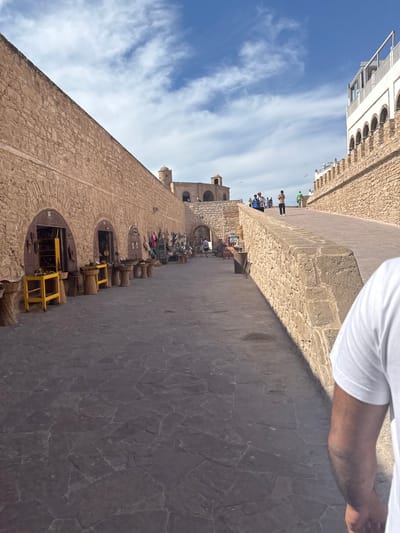The image captures a scene within the historic ramparts of Essaouira, Morocco. The viewpoint is from a cobblestone pathway, looking towards a long, fortified stone wall on the left, lined with arched openings that serve as shops. These shops display various goods, including baskets, wooden furniture, and what appear to be handmade crafts. In front of the shops, woven baskets and yellow wooden tables are arranged. Further down the pathway, more arched structures and stone towers are visible, consistent with the architecture of Essaouira's medina. On the right side of the image, a higher rampart wall made of rough-hewn stones rises, with a modern building with white facade and arched windows visible at its top. Several people are walking along the ramparts, appearing to be tourists. The sky is blue with scattered white clouds. In the foreground, the shoulder and arm of a person wearing a white t-shirt are visible, indicating the photographer's perspective. The overall scene depicts a bustling historic site with commercial activity.