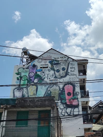 This image captures an urban scene in Thành phố Đà Lạt, Vietnam, primarily featuring the side of a multi-story building. The building has a white facade and a reddish-brown tiled gable roof. A large, colorful mural covers a significant portion of the white wall, advertising beverages. The mural depicts various drinks, including cups labeled "Green Tea Milk Tea," "Taro Milk Tea," "Grape Tea," "Peach Tea," "Longan Tea," and "Passion Tea." Several illustrations of bubble milk tea with dark pearls are visible, alongside a pink blended drink with strawberries. Text on the mural includes "Say Hi... Bubble Milk Tea," "fresh," "Coffee," and Vietnamese phrases such as "Thức Uống Dinh Dưỡng" (Nutritious Drinks), "Trà Sữa Trân Châu" (Bubble Milk Tea), and "Trà Chanh" (Lemon Tea).

A metallic cylindrical water tank, supported by a black metal frame, is mounted on the roof of the building, positioned to the left of the gable. Multiple dark electrical or communication wires run horizontally across the foreground, partially obscuring the building. On the right side of the main building, two balconies with black wrought-iron railings are visible. Below the main building, to the left, is an older, lower structure with a grey exterior and two closed green wooden doors or shutters. A section of another building with a dark roof is partially visible on the far right. The sky above is bright blue with scattered white cumulus clouds, indicating daylight conditions. No people are visible in the scene.