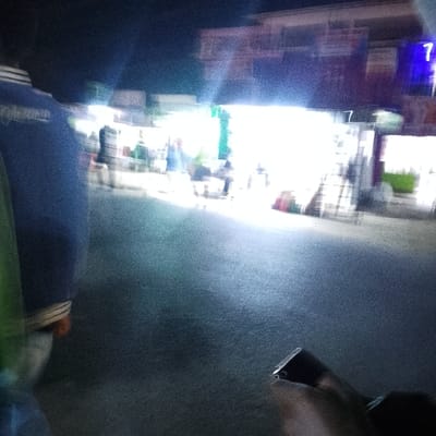 A blurry nighttime scene depicts a crowded street with vendors and people. Bright lights obscure details of the stalls. In the foreground, a person wears a blue jacket with white stripes, partially obscuring the view. Below, what appears to be a hand and a dark object rest on a vehicle handlebar. A building with a sign showing the number 7 is faintly visible in the background. The overall image quality is poor due to motion blur and low light conditions.
