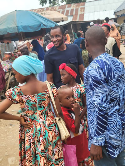 The image depicts a crowded scene, possibly a marketplace in Madalla, Nigeria. People of various ages are present. A woman with a light blue head covering and a colorful patterned dress stands near the lower edge of the frame. A man in a dark blue shirt is smiling and looking at the camera. Two children, one wearing a red headband and the other in a red shirt, are in the scene. Another man is wearing a blue patterned shirt. A blue tent-like structure is visible in the background, along with various stalls and other individuals.