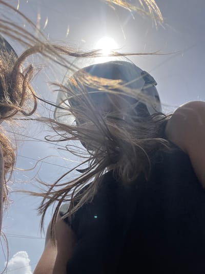 A low-angle, close-up shot captures a person wearing a helmet and a black shirt, with their hair blowing in the wind against a bright sky with the sun visible. The person's left arm is bent and raised, with their hand near their face. The image appears to have been taken in Goviyapana, Sri Lanka, suggested by the presence of overhead power lines and a distant cumulus cloud, characteristic of the tropical climate. The overall lighting suggests it is daytime.