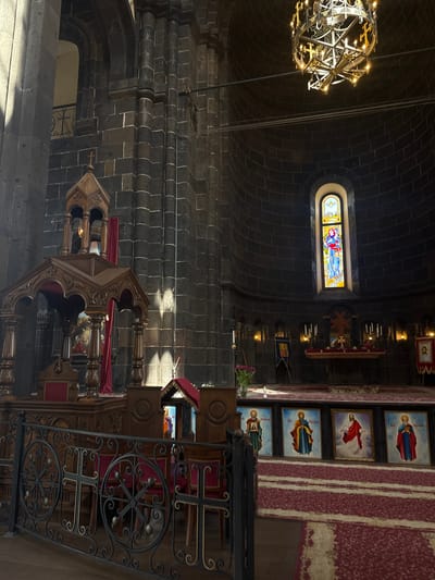 This image depicts the interior of the Holy Mother of God Cathedral in Gyumri, Armenia. The cathedral features dark stone walls and a high, domed ceiling. A large, ornate chandelier hangs from the ceiling. In the center of the image, a wooden pulpit or ambo with intricate carvings stands on a red carpeted floor. Behind it, a dark wooden railing with ornate metalwork separates the congregational area from the altar.

The altar area is adorned with religious icons, candlesticks, and banners. A stained-glass window featuring religious figures is visible above the altar. Sunlight streams through the window and illuminates parts of the stone walls, creating dramatic light and shadow effects. The overall atmosphere is solemn and reverent, characteristic of a place of worship.