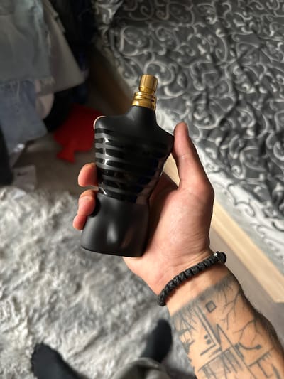 A person's hand holds a black, torso-shaped perfume bottle with a gold cap. The bottle has horizontal black stripes. The hand also wears a black braided bracelet. The person has tattoos on their forearm. The background includes a gray, patterned bedspread, a fluffy gray rug, and some clothing items in the upper left corner. The scene appears to be indoors, likely in a bedroom. There is no visible indication of the city of Kazanlak, Bulgaria in this image.