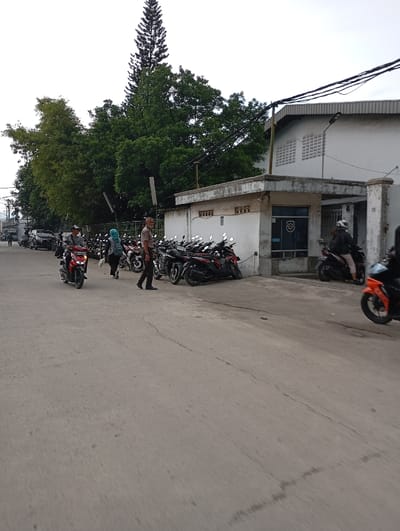 The scene shows a street in Katapang, Indonesia (latitude: -6.986, longitude: 107.555). A group of people are standing near a row of parked motorcycles. A man in a brown shirt is walking towards the camera, while a woman in a blue headscarf is standing next to a motorcycle. In the background, a white building with a grey roof is visible. Several power lines are strung across the street, and the building appears to be a business or industrial facility. The image was taken from a moving vehicle. The asphalt road is in good condition, with some minor cracks. There is green foliage and a tall tree on the left side of the image. The sky is a light grey. The sun is not visible. The image is taken at a slight angle.