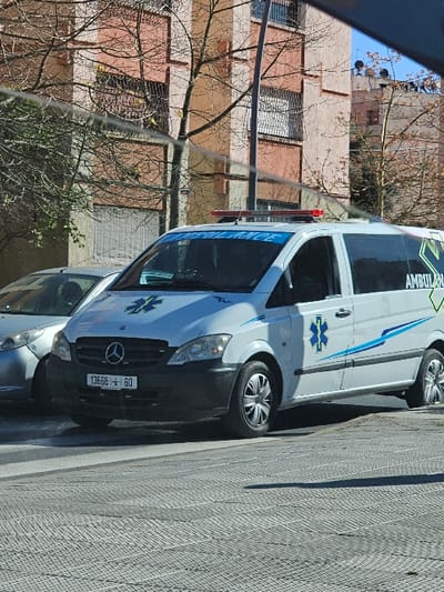 The scene depicts a white Mercedes-Benz van ambulance, identified by its blue "Star of Life" emblems and "AMBULANCE" text on the windshield and side, parked or stationary on a paved surface in Salé, Morocco. The vehicle displays a Moroccan license plate reading "13668 | 60" and features a red emergency light bar on its roof. A silver passenger car is partially visible behind and to the left of the ambulance. In the background, a multi-story residential building with a peach-colored facade, multiple windows, and balconies with dark metal railings is visible. Deciduous trees with bare branches are situated between the vehicles and the building. The foreground consists of light-colored, textured paving stones. The sky is clear blue, indicating bright daylight conditions. No individuals or specific actions are discernible.