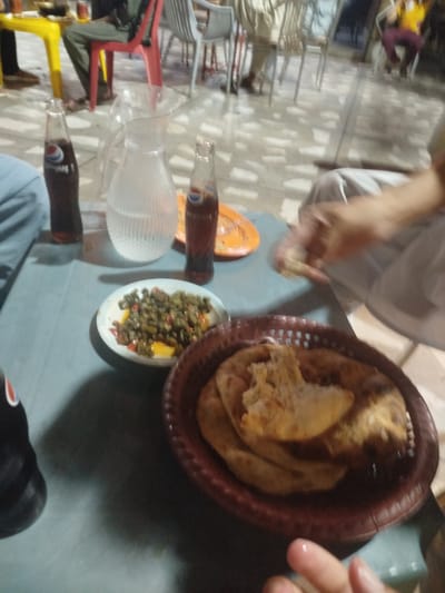The image depicts an outdoor dining scene, likely at a restaurant in Peshawar City Tehsil, Pakistan, given the context provided. The foreground is dominated by a dark grey table where a meal is laid out. On the table, there are two glass bottles of Pepsi, a clear glass pitcher filled with water, a small white plate containing a green vegetable dish with some red and yellow pieces, and a woven brown basket filled with pieces of flatbread. A hand, with a finger that appears to be wet or oily, is reaching towards the bread from the right side of the frame.
In the background, several people are visible sitting at tables. To the left, a person in dark clothing is seated on a bright yellow stool. Behind them, a person in green trousers and a dark top is seated on a red chair. Further in the background, several other individuals are seated on chairs around tables, with some appearing to be enjoying their meals. The environment suggests a casual, open-air dining setting, possibly with a patterned tiled floor. The lighting is subdued, contributing to a somewhat dimly lit atmosphere.
