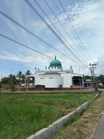 A mosque with a green dome is situated in Meukokuthang, Indonesia. The building is white with green trim and multiple smaller green domes. It is surrounded by a low stone wall and a grassy area. In the foreground, there is a field of tall green grass and a concrete drainage ditch. In the background, palm trees and other vegetation are visible, along with power lines stretching across the frame. A utility pole with wires is also present. A small building and a black car are visible to the right of the mosque. The sky is blue with scattered clouds.