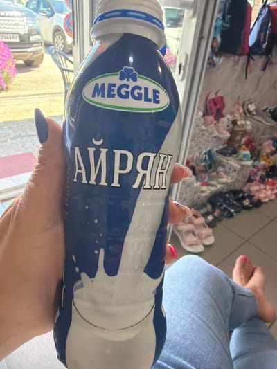 In Ravno pole, Bulgaria, a person is holding a bottle of "AYRAH" branded drink. The bottle has a blue and white label with the brand "MEGGLE" in green. The person's hand, with dark blue nail polish, is visible on the left side of the bottle. In the background, a car is parked outside what appears to be a shop, with more bottles or containers stacked on shelves and a selection of footwear, including sandals and sneakers. A person's legs, wearing blue jeans, are visible in the lower right corner of the frame.