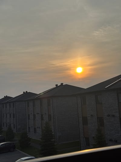 The image captures a dusk scene in Gatineau, Canada, featuring a row of identical brick apartment buildings with dark sloped roofs. The sun, a bright yellow orb, is partially obscured by a hazy, pale orange sky filled with thin clouds. The apartment buildings, constructed with light gray bricks and accented with vertical bands of brown siding, are positioned in a linear arrangement. Several windows are visible on the facades of the buildings. Small evergreen trees are planted in front of the buildings. In the foreground, a parking lot is visible with a dark-colored SUV and the edge of a white car. The overall environment suggests a residential area under a softly lit sky.