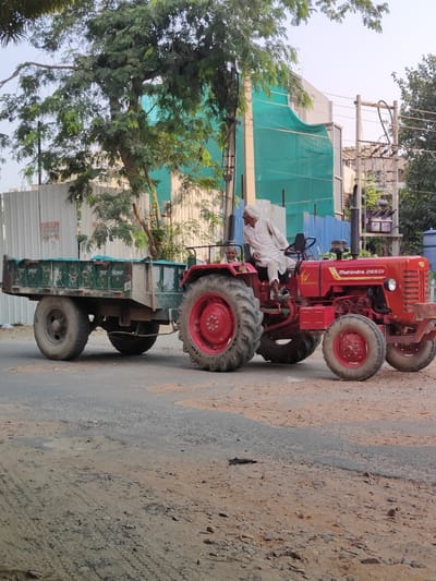 A red Mahindra 265 DI tractor is driving on a dusty road in Gurgaon, India, pulling a green trailer. A man wearing a white turban and traditional Indian attire is driving the tractor. Another man is visible in the passenger seat. The tractor and trailer are on the right side of the road, with a construction site in the background, featuring a green mesh covering and scaffolding. Trees and power lines are also visible in the surrounding environment. The road surface is a mix of asphalt and loose dirt and gravel. The overall scene depicts a typical day in a developing urban area.
