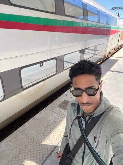A male individual is captured in a selfie at the Tanger Ville railway station in Tanger, Morocco, standing on a light-colored concrete platform beside a stationary Al-Boraq high-speed train. The man, approximately 30-40 years old, has dark, curly hair, a thick mustache, and is wearing dark sunglasses with tortoiseshell frames. He is dressed in a light grey or sage green hooded jacket with a black shoulder bag featuring a strap with red and teal stripes. The train, a modern double-decker model, is predominantly white with horizontal red and green stripes running along its side. Sunlight casts distinct shadows on the platform, indicating clear weather. Overhead catenary lines are visible above the train in the background against a clear blue sky.