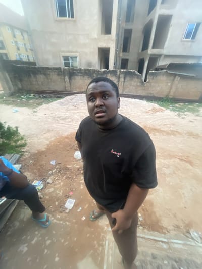 The image displays a full-body shot of a man taken from a slightly elevated perspective, with the original photo appearing to be rotated 90 degrees counter-clockwise.
The primary subject is a dark-skinned man with short dark hair and a beard/stubble. He is wearing a plain black short-sleeved t-shirt with a small, indistinct logo on the left chest area, which appears to be three tiny, stacked shapes, possibly hearts, in a light color (like red or pink). He is also wearing dark-colored trousers and light blue flip-flops. The man is looking directly towards the camera with a surprised or attentive expression, his mouth slightly open. He is standing with his body angled slightly to the side, leaning forward. His right hand is visible resting on his right thigh, while his left arm is mostly out of frame or obscured.
The setting is outdoors on what appears to be an unpaved ground surface composed of dirt, sand, and possibly some dried mud or wet patches. Scattered pieces of refuse, such as plastic bags and wrappers, are visible on the ground around the man.
In the background, several multi-story buildings are visible. These structures appear to be unfinished or under construction, characterized by exposed concrete or cinder block walls that have been rendered but not yet painted. Many window openings are visible, some with glass installed. The architecture suggests a residential or commercial area. A concrete wall separates the foreground ground from the buildings in parts.
In the lower right corner of the image (when oriented correctly), a portion of another person's legs and feet is visible, also wearing dark trousers and light blue flip-flops, suggesting another individual is nearby, possibly seated or partially out of frame.
The lighting indicates it is daytime, with good natural illumination, suggesting it might be late morning or early afternoon. There are no strong shadows, creating an evenly lit scene. No specific text is clearly legible on any objects or surfaces within the image, except for the small, indistinct logo on the man's shirt. The overall scene conveys an informal, outdoor setting in what appears to be a developing or less developed urban or semi-urban environment.