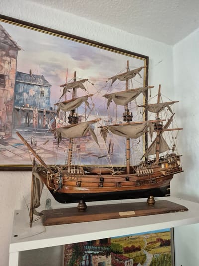 A wooden model of a sailing ship is displayed on a white shelf in front of a framed painting of a harbor scene. The ship model is detailed, with multiple masts, sails, rigging, and cannons. The painting depicts buildings along a waterfront with a cloudy sky. The overall scene is indoors, with a white textured wall in the background. The location is identified as Trier, Germany.