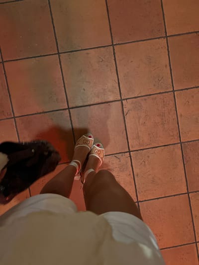 A person is standing on a tiled floor in a location in Miami Beach, Florida, at the coordinates 25.769, -80.135. The floor is made of large, square, terracotta tiles with a red hue. The person is wearing white shorts and beige sandals with a Gucci logo on them, and they have red nail polish. The person's lower legs, feet, and part of their torso are visible in the image. The person is holding a black purse, which is partially visible in the lower left corner of the image. The lighting is dim, and the person is casting a shadow on the floor. The person's feet are facing forward and their legs are slightly spread apart. The person appears to be standing still. There is no one else present.