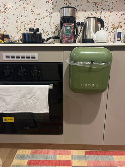 The image depicts a kitchen setting, likely in an apartment or house, given the countertop and cabinet setup. The location in Miraflores, Peru, is not ascertainable from the visual data. On the left, there is a countertop with a speckled pattern, holding a coffee maker, an electric kettle with a teapot on top, and a bottle of spa lotion. A green UFO RU branded breadbox sits on top of a beige cabinet unit with a built-in oven. The oven door is black and has a white towel hanging from it. A ventilation grill is located to the left of the oven door. To the right of the cabinet unit, there is a vertically oriented rug with alternating bands of red, blue, and yellow colors.