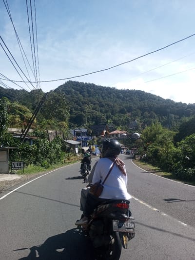 The image depicts an outdoor scene on a clear, sunny day, captured from the perspective of a person riding a motorcycle on a paved road. The environment transitions from a winding road lined with vegetation to a mountainous backdrop.
In the immediate foreground, the back of a person riding a dark-colored scooter or motorcycle is visible. The rider wears a dark helmet, a white long-sleeved shirt, light-colored pants, and light-colored sandals. A reddish-brown scarf or fabric is draped over their shoulders, and a brown shoulder bag with a red strap is worn across the body. The motorcycle features a red tail light assembly and a rear license plate displaying "F 4837 FGH". Long shadows from the motorcycle and rider are cast onto the road to the left.
Ahead of the foreground vehicle, two other motorcycles are visible, traveling in the same direction on the two-lane road. The road surface is dark grey asphalt with a solid white line marking the right edge and a broken white center line. It curves gently to the right in the mid-distance.
The left side of the road features a white sign with "PENGINAPAN VILLA KAMAR" written in black capital letters. Behind it, dense green foliage, trees, and utility poles with numerous overhead wires are present. Further back, several buildings are visible, including a prominent blue structure with the word "POLISI" (Police) written in white capital letters on its facade.
The right side of the road is bordered by dense green vegetation. In the background, a large, heavily forested mountain rises, covered in dark green trees under a light blue sky with scattered thin white clouds. Scattered residential or commercial buildings are visible on the slopes and in the valley below the mountain, including some with reddish-brown roofs and at least two distinctive white-domed structures.
While the prompt specifies Ka Thaung (Upper), Myanmar, visual evidence within the image, such as a building sign displaying "POLISI" and a motorcycle license plate format "F 4837 FGH," suggests a location in Indonesia.