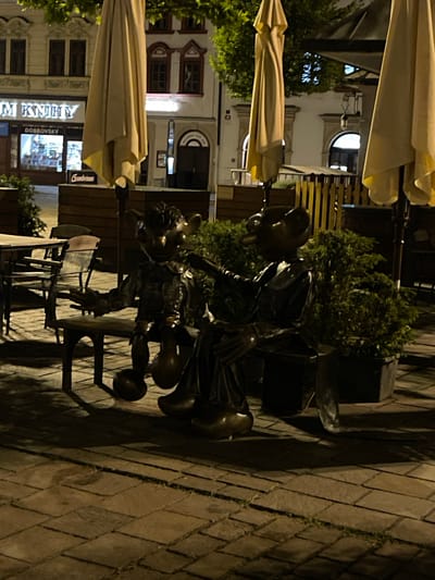 This image depicts a nighttime scene on a cobbled street in Pilsen, Czechia, featuring two bronze statues of characters resembling mice or puppets sitting on a bench. The statues are positioned outdoors, with several large, closed umbrellas and greenery in the background. One statue, on the left, has its arms outstretched, while the other, on the right, appears to be leaning towards the first. In the background, a building with illuminated windows and signage is visible, suggesting a commercial establishment. The lighting suggests it is evening or night, with artificial lights illuminating the area and casting shadows. The overall atmosphere is quiet and still.