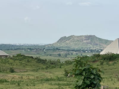 This is a landscape photograph taken in Gidan Waziri, Nigeria. The scene depicts a rural or semi-rural environment with rolling green hills covered in grass and scattered trees. In the foreground, there is a large leafy green bush. To the left, a portion of a building with a slanted gray roof is visible.

In the middle ground, the landscape continues with more green vegetation and some scattered structures. A prominent feature in the middle distance is a large, barren, rocky hill with sparse vegetation. Beyond this hill, a distant city or town is visible, characterized by a dense collection of buildings.

On the right side of the image, a large, light-colored, pyramid-shaped structure with a metallic roof stands out against the background. The sky is overcast, suggesting a cloudy or hazy day. The overall impression is one of a vast, open landscape with signs of both natural terrain and human development. There is no discernible activity or event taking place, and no visible text. The time of day appears to be daytime, but the weather conditions obscure any direct sunlight.