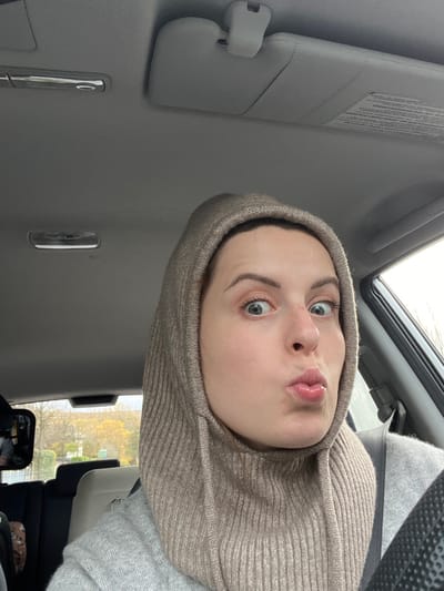 A light-skinned female individual is depicted in the driver's seat of a vehicle in Staudernheim, Germany. She is centered in the frame, wearing a light beige ribbed balaclava that covers her head and neck, and a grey sweater. Her eyes are wide open, and her lips are puckered in an exaggerated facial expression. The interior of the vehicle features a grey headliner with two overhead light fixtures and a grey sun visor on the passenger side. A black steering wheel is partially visible on the bottom right, and a seatbelt crosses her right shoulder. A rearview mirror on the left reflects a blurred object, consistent with a child car seat. Through the vehicle windows, an overcast sky and autumnal trees with yellow foliage are visible in the background.