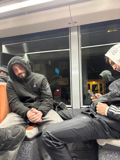 Two men are on a train at night. One man, wearing a black jacket and a hooded shirt, rests against the window. His eyes are closed, and his hands are clasped. The other man is standing beside him, facing away, wearing black Adidas track pants and jacket, and looking at his phone. The train's window shows reflections of buildings outside.