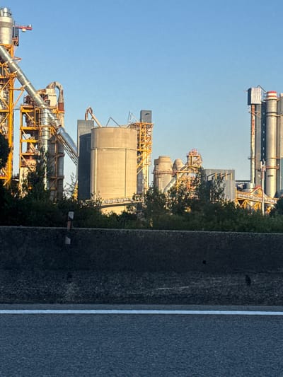 The image shows an industrial complex in Castanheira do Ribatejo e Cachoeiras, Portugal, likely a cement factory or similar facility. The complex features tall cylindrical structures, conveyor belts, and a yellow metal framework supporting various industrial components. The sky is clear and blue, indicating daytime. In the foreground, a concrete barrier and a section of asphalt road with a white line are visible, suggesting the image was taken from a roadside perspective. Lush green trees and foliage partially obscure the base of the industrial structures. The overall scene depicts an operational industrial zone within a natural landscape.