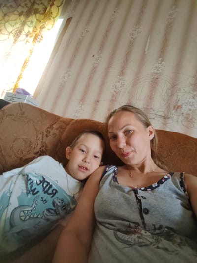 In this medium shot, a woman and a young boy are resting on a brown couch, likely indoors. The woman, positioned on the right, is looking towards the camera with a slight smile. She has blonde hair and is wearing a gray tank top with a floral trim. A delicate silver necklace adorns her neck.
To her left, the boy is also looking towards the camera, with his head tilted slightly. He is wearing a light blue long-sleeved shirt with a graphic print of a shark or dinosaur. The shirt has text that appears to be in Russian, with some letters visible such as "RIDA" and possibly "M." The couch has a textured pattern, and behind them, the wall is covered in patterned wallpaper with vertical stripes and floral motifs in shades of cream and pink.
To the far left, a window is visible, partially covered by a sheer yellow curtain with a floral design. Sunlight streams through the window, suggesting it is daytime. The overall atmosphere seems relaxed and casual, indicating a moment of shared downtime for the two individuals. The setting appears to be a home interior in Чухлэм, Russia.