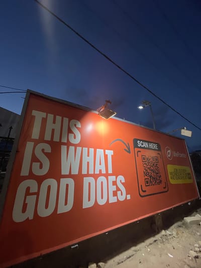 The image shows a sign for LifePointe Church in Ilado, Nigeria. The sign is red and prominently displays the text "THIS IS WHAT GOD DOES." It also features a QR code with the instruction "SCAN HERE," and mentions service times: "JOIN US EVERY WEDNESDAY AT 6:18PM AND SUNDAYS AT 10AM." The photo was taken at dusk. A utility pole and wires are also visible.