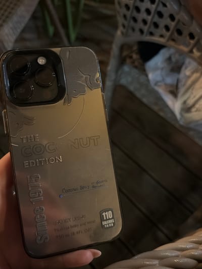 A smartphone with a decorative case resembling a vintage coconut beverage is held in a hand. The case features text "THE COCONUT EDITION", "Since 1975," and nutritional information, including "110 CALORIES". The phone has a multi-lens camera array. The background is out of focus, showing indistinct textures and patterns. No location details, specifically Elhovo, Bulgaria, are evident in the image.