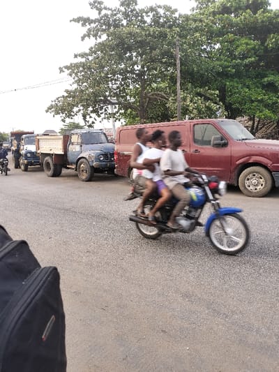 The image captures a dynamic street scene during daylight, likely in an urban or semi-urban setting, characterized by traffic and a mix of vehicles.
In the immediate foreground, a blurry black object, possibly a backpack or bag, occupies the lower-left corner, suggesting the photo was taken from a pedestrian's perspective or from another moving vehicle. The road surface visible is asphalt, showing some wear.
The central focus of the image is a blue motorcycle in motion, evidenced by the significant motion blur on its wheels and the three individuals riding it. The driver, positioned at the front, wears a light-colored t-shirt and what appears to be light brown or beige trousers. Immediately behind the driver is a person wearing a white top and purple shorts. The third person is seated at the very back, wearing a dark top and dark shorts. Their faces are indistinct due to the motion blur. The motorcycle itself is a standard blue model, possibly a commuter bike, with silver-colored rims.
To the right of the motorcycle, a dark red or maroon panel van is visible, parked or moving slowly. Its side door appears to be of the sliding variety.
Further into the midground, to the left of the motorcycle, a line of heavy-duty trucks stretches down the road. The most prominent is an older model blue truck with an open flatbed or dump bed, exhibiting visible signs of wear, dents, and rust, suggesting considerable use. Its tires appear robust and well-worn. Behind this blue truck, another truck of similar design, but a lighter color (possibly white or light blue), is also part of the queue. Further down the road, another smaller motorcycle with a rider is visible near the line of trucks.
The background is dominated by a dense canopy of lush green trees, particularly on the right side and across the upper portion of the frame, indicating a rich natural environment. A utility pole stands vertically among the trees on the right. The sky, visible in the upper-left, appears bright and hazy or overcast, typical of daylight conditions.
The overall scene conveys a sense of everyday life and traffic flow, with the overloaded motorcycle being a notable detail. No discernible text is visible on any of the vehicles or in the environment.