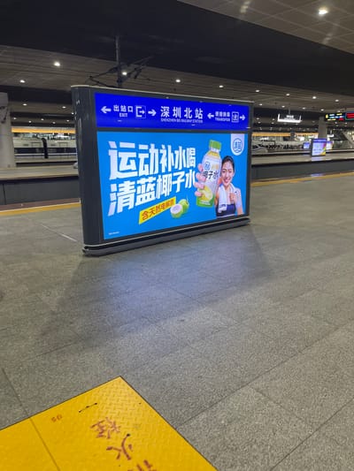 This is a medium-shot, eye-level, outdoor photograph taken at a train station in Shenzhen's Longhua District, China. The main subject is a large, illuminated advertisement displayed on a freestanding digital billboard. The billboard features Chinese text and an image of a woman holding a bottle of "Qinglan Coconut Water." The ad promotes the beverage as a source of natural electrolytes.
Above the advertisement, a blue directional sign indicates "EXIT" with an arrow pointing left and "SHENZHEN BEI RAILWAY STATION" with an arrow pointing right. To the right of the station name, another directional sign indicates "TRANSFER" with arrows pointing left and right. The train station platform is visible in the background, with a train parked at the platform. The floor is paved with grey granite tiles and a yellow diamond-plate safety zone is present in the foreground. The lighting suggests it is daytime.