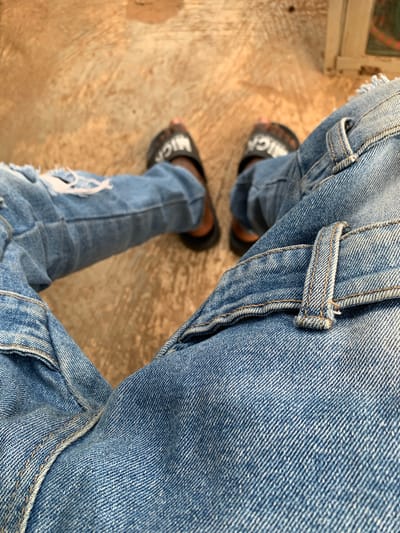 The image presents a high-angle, close-up view of a person's lower body, focusing on their legs and feet. The subject is wearing light blue, distressed denim jeans with visible rips on the left leg, particularly around the knee area. The jeans feature prominent yellowish-brown stitching along seams and a belt loop, highlighting the garment's construction. The denim fabric displays a clear twill weave pattern.
The person is wearing dark-colored slide sandals. On the left sandal, part of a white word, "BALENCI", is visible. On the right sandal, another part of a white word, "MICA", is visible, indicating a brand name, likely "BALENCIAGA".
The background is a light brown or tan ground surface, which appears textured and somewhat worn, possibly concrete or a similar hard outdoor surface. The lighting suggests it is daytime. The overall scene captures a casual moment, with the perspective looking down at the feet. The location context is Kabba, Nigeria.
