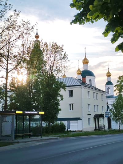 The image captures a street scene in Chelganovo, Russia, featuring a prominent religious building with multiple gold-domed spires. The building, painted a light yellow, is partially obscured by trees and sunlight. In the foreground, a bus stop shelter with a digital display is visible. The road in front of the bus stop is asphalt. The sky above is partly cloudy, with the sun casting a warm glow through the trees. There are no people visible in the image.