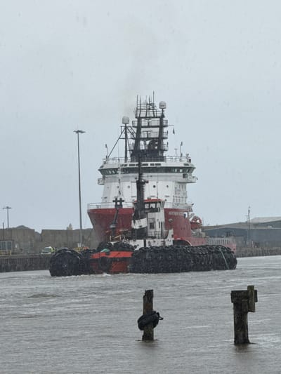 The image shows a red and white tugboat, identified by the text "NORTHERN VICTORY" on its side, navigating through choppy, gray water under a cloudy sky. The tugboat is equipped with multiple radar dishes and antennas on its upper structure. Smoke or steam emanates from the top of the vessel. The foreground of the image features two wooden posts in the water, each wrapped with black fenders. In the background, a dock or pier area is visible with several buildings and what appears to be industrial infrastructure. There are also some vehicles, including what looks like a white van, present on the dock. The overall atmosphere is overcast, suggesting inclement weather, with visible rain or spray in the air. The scene is set in the port area of Great Yarmouth, United Kingdom, with the waters of the River Yare or the harbor being depicted.