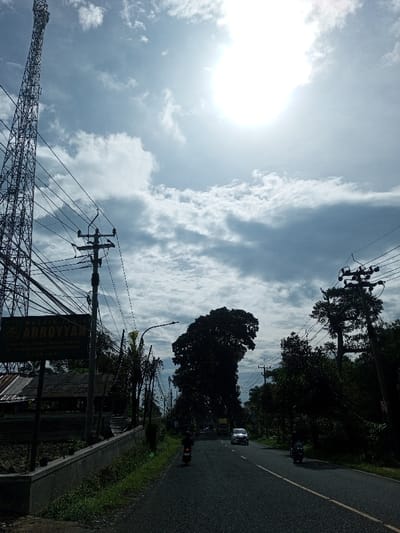 The image depicts a daytime outdoor scene along a paved road in Domphoog_Dungkarchhoeling, Bhutan. The bright sun is visible in the upper right quadrant of a partially cloudy blue sky, causing significant glare and backlighting.
In the foreground, a two-lane asphalt road extends into the distance. Two motorcycles, each with a single rider, are traveling away from the viewer in the right lane; one is closer in the foreground, and the other is further down the road. A white sedan is visible beyond the second motorcycle, also in the right lane, followed by a light-colored truck further in the distance.
On the left side of the scene, a tall, metallic lattice communications tower stands prominently. Below it, a utility pole densely wired with black cables rises next to a concrete retaining wall. Behind the wall, a sign with the prominent text "ARROYYAN" is mounted on a low-rise structure featuring a corrugated metal roof. Roadside vegetation, including grass and weeds, grows along the asphalt edge.
The right side of the road is bordered by multiple utility poles with overhead wires and dense green foliage, including several large trees. A particularly large, dark-foliaged tree stands prominently in the midground, silhouetted against the bright sky. The road continues into a treelined background.