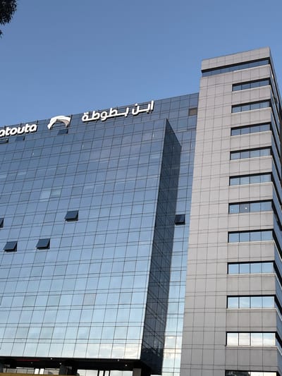 The image depicts a tall, modern building in Tanger (طنجة), Morocco. The building has a glass facade, with many windows reflecting the blue sky. A portion of the building has a gray exterior with black-framed windows. The name "Ibn Battouta" (ابن بطوطة) and an associated logo are visible on the building's exterior. Several windows are partially open. The overall perspective is tilted, showing the building from a low angle.