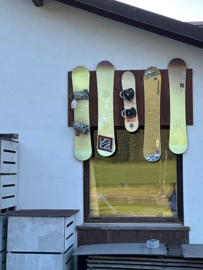 The image shows five snowboards mounted vertically on a white stucco wall in front of a building with a dark-brown roof overhang. The snowboards are arranged from left to right, with the first and last boards being light yellow, the second and fourth boards being a lighter shade of yellow, and the middle board being a golden-yellow color. The snowboards have bindings attached, and some have graphic designs or logos. A large window is visible below the snowboards, reflecting a green field. To the left of the wall are white slatted storage units. In the foreground, there are stacks of wooden planks and a small grey stone cube on top of a plank. The scene appears to be outdoors, possibly at a ski resort or a residence in Le Brassus, Switzerland, given the presence of snowboards and the architectural style.